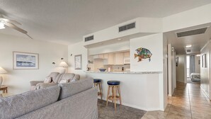 A modern living room with a sofa, a dining area with stools, and a kitchen with a white countertop. - Sea Gull #704: Beachfront 2 Bedroom 2 Bathroom With Spectacular Views and 24 Hour Management (Port Aransas)