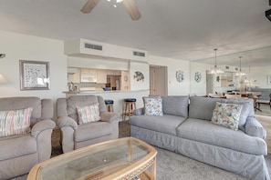 A modern living room with a grey sofa, armchairs, and a glass coffee table. A kitchen with white cabinets and a dining area with a table and chairs are visible in the background.