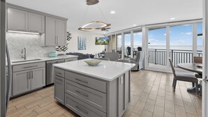 A modern kitchen with grey cabinetry, a central island, and a dining area with a view. - Sea Gull #906: Beachfront 2 Bedroom 2 Bathroom With Spectacular Views and 24 Hour Management (Port Aransas)