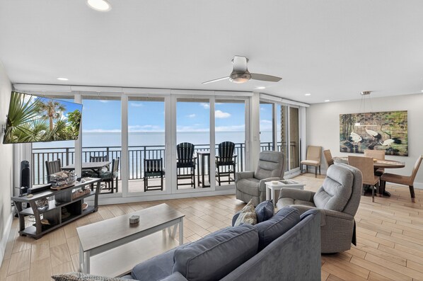 A spacious living area with a balcony, large windows, a ceiling fan, and a flat-screen TV.