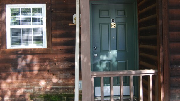 Une cabane en bois avec une porte verte, une fenêtre avec des stores et une véranda dotée d’une balustrade.