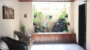 Una habitación con un banco de madera, sillas de mimbre y un pequeño jardín interior con plantas y una formación rocosa.