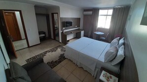 Una habitación de hotel con cama, un escritorio pequeño, televisor, sofá y ventana con cortinas.