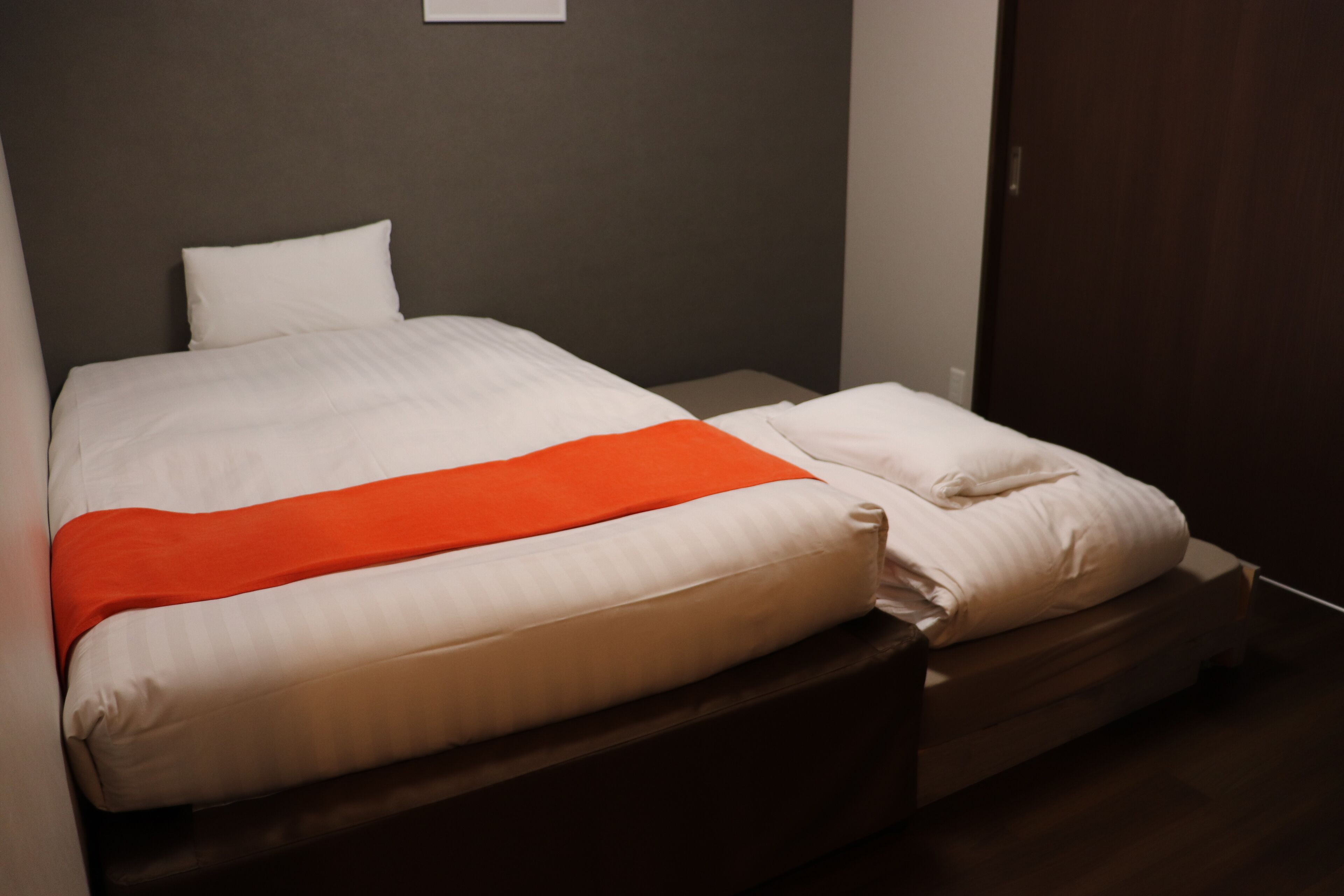A neatly made bed with white bedding and a red accent, a wooden headboard, and a dark brown door in a hotel room.