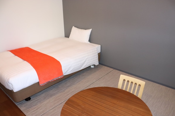 Una cama individual con colchón blanco y manta naranja, una silla de madera y una pequeña mesa redonda en una habitación con paredes grises y suelo de madera.