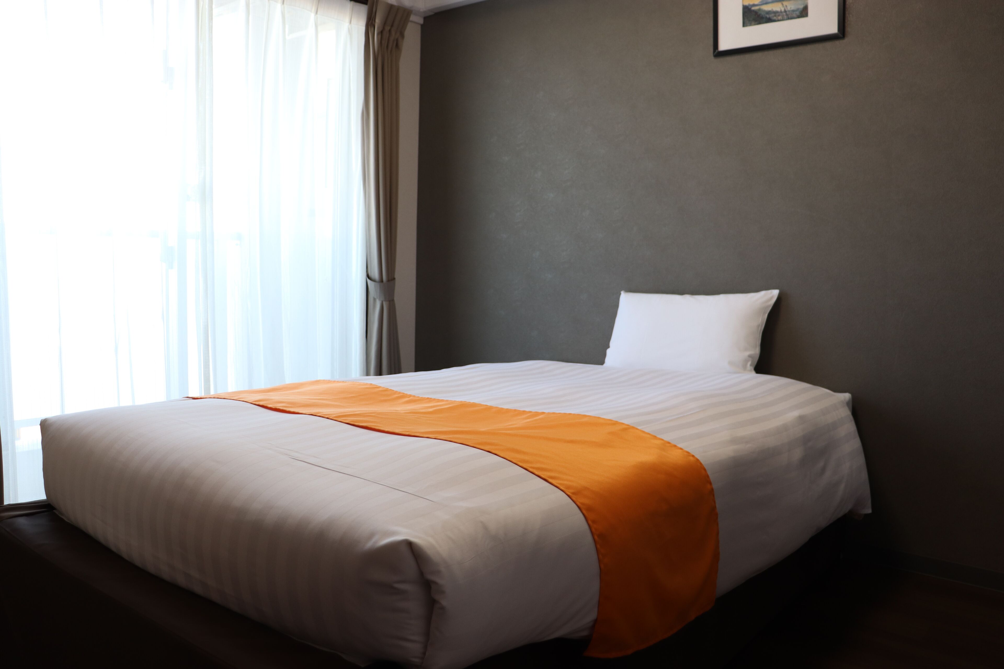Un lit bien fait, recouvert d’une couverture blanche et orange, avec un oreiller blanc et un tableau encadré accroché au mur.