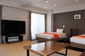 A hotel room with a flat-screen TV, two beds with orange blankets, a wooden table, and a window with curtains. - ApartmentHotel ecott (Kagoshima)