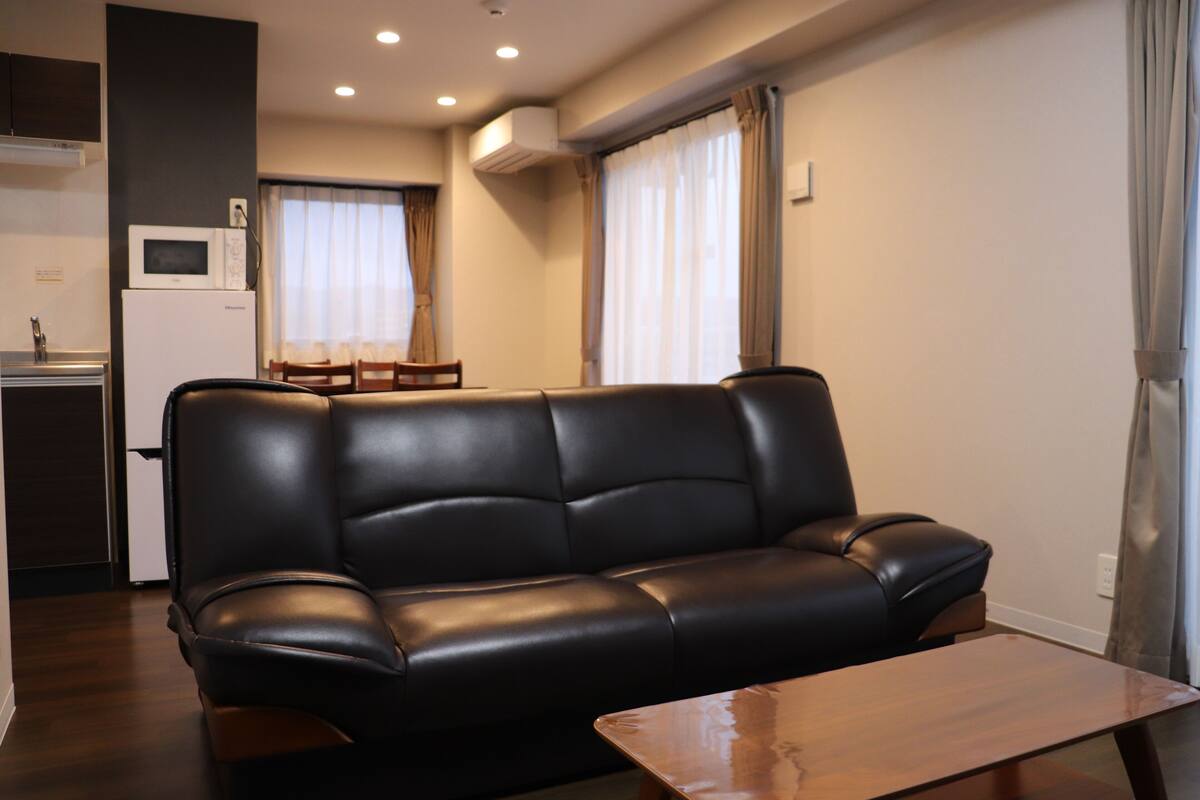 A modern living room with a black leather sofa, a wooden coffee table, and a kitchen area in the background.