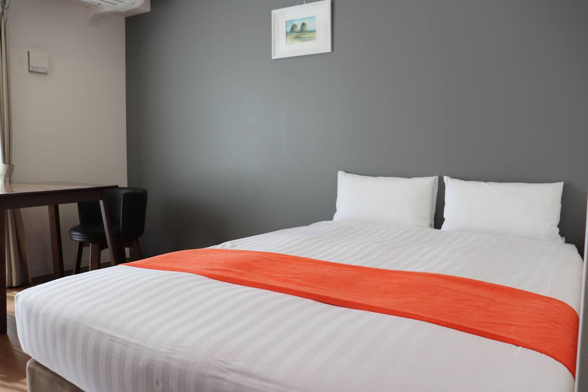 A neatly made bed with white bedding and an orange accent, a desk with a chair, and a framed picture on the wall.