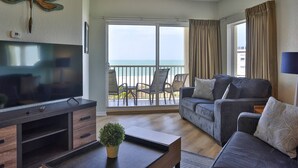 A living room with a TV, sofa, and a balcony with a view of the beach. - Family Vacay-Beach Days, Pool Time, Hot Tub, Spectacular Views. Beachfront Pools and Bar, Cafe, Gym (Treasure Island)