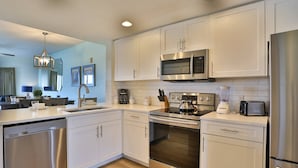 A modern kitchen with stainless steel appliances, white cabinetry, and a dining area with a table and chairs. - Family Vacay-Beach Days, Pool Time, Hot Tub, Spectacular Views. Beachfront Pools and Bar, Cafe, Gym (Treasure Island)