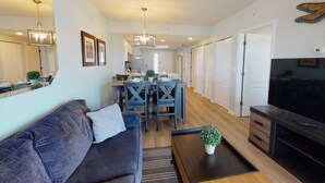 A modern living room with a sofa, coffee table, TV, and dining area with chairs and a table. - Family Vacay-Beach Days, Pool Time, Hot Tub, Spectacular Views. Beachfront Pools and Bar, Cafe, Gym (Treasure Island)