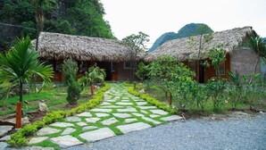 A courtyard with thatched-roof huts, a stone pathway, and lush greenery. - Trang An Mountain House - Bungalow Deluxe with garden view - Ninh Binh (Ninh Binh)