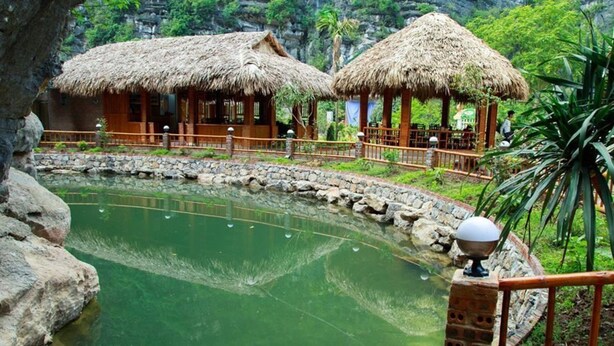 A rustic lodge with thatched-roof huts, a stone-walled pond, and a wooden deck. - Trang An Mountain House - Bungalow Deluxe with garden view - Ninh Binh (Ninh Binh)
