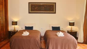 A hotel room with two beds, each with a brown bedspread and a white towel. There are two bedside tables with lamps, a framed picture on the wall, and a wooden door on the left.