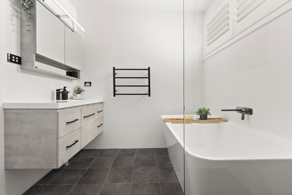 A modern bathroom with a white vanity, a large mirror, a bathtub, and a towel rack. - Outstanding ocean views (Mollymook Beach)