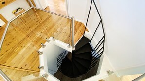 En spiraltrappa med glasväggar och metallräcken, som leder upp till en liten plattform med trägolv och en vit vägg.