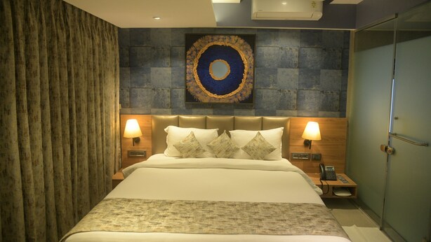 A hotel room with a large bed, two bedside lamps, a wall-mounted artwork, and a ceiling fan. - Hotel Bindra Supremacy (Mumbai)