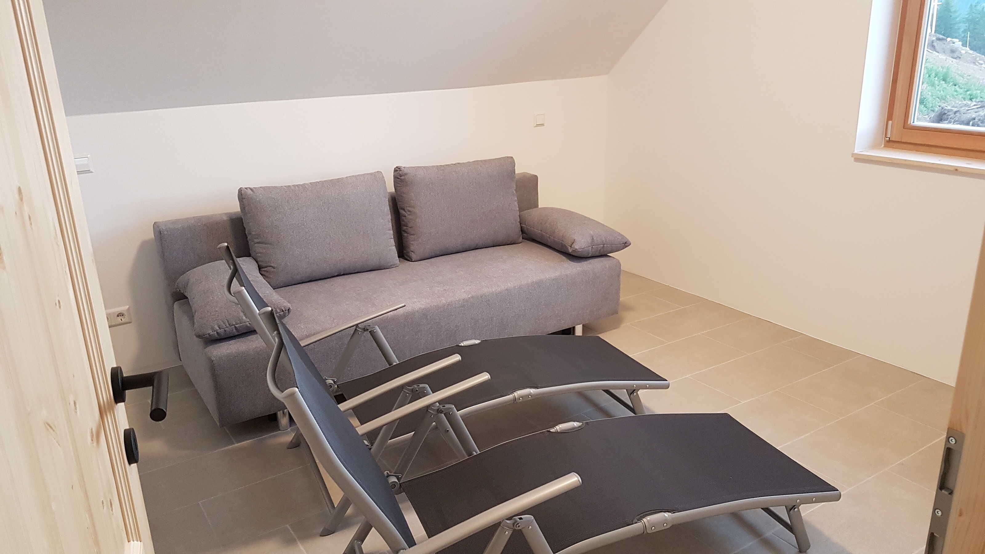 A room with a grey sofa, a foldable table, and a window with a view of trees.