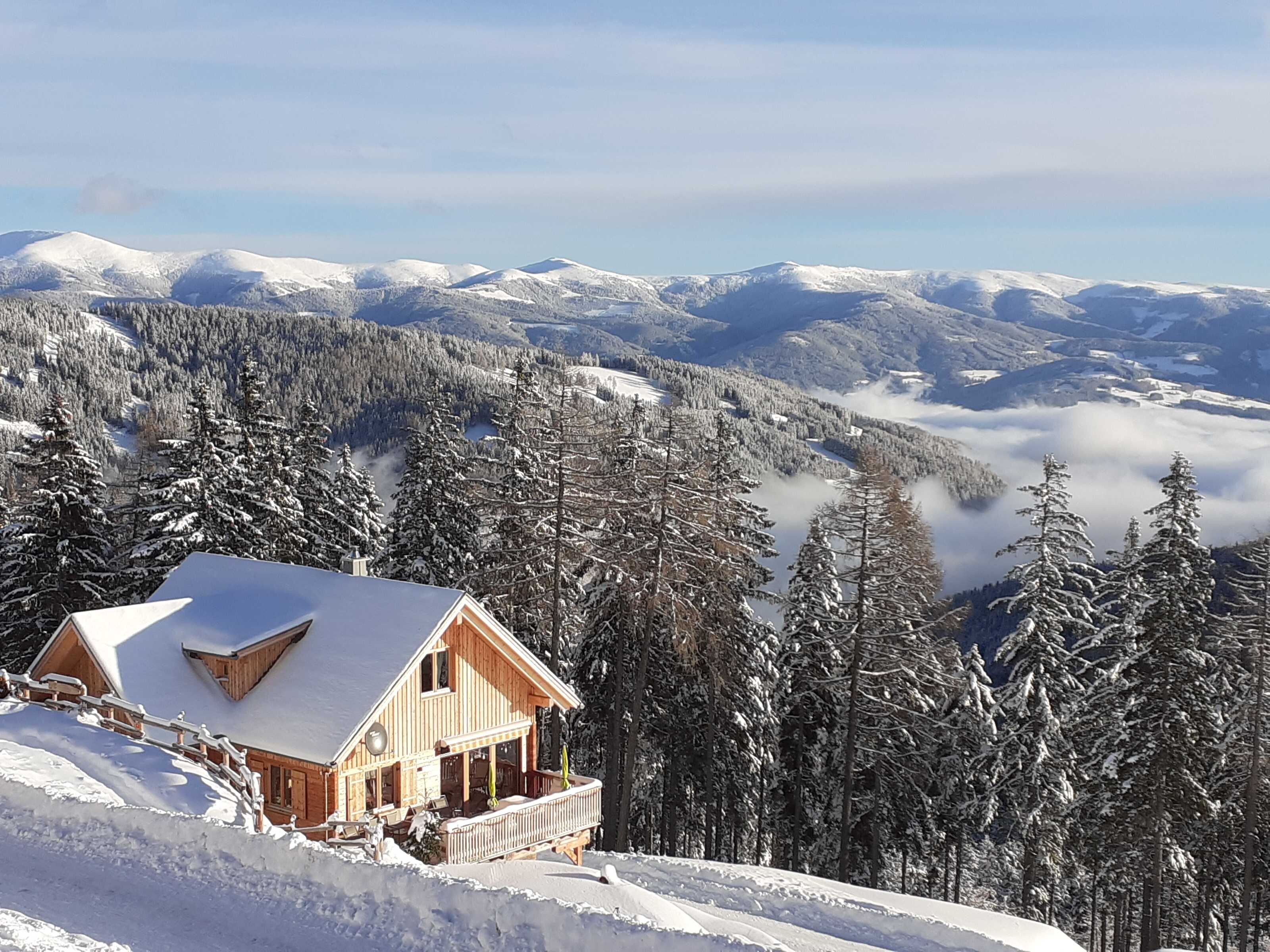 Una cabana de fusta amb teulada inclinada, envoltada per arbres coberts de neu i muntanyes al fons.