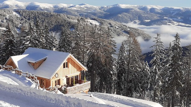 Une cabane en bois, dotée d’un toit en pente, entourée d’arbres couverts de neige et avec des montagnes en arrière-plan.