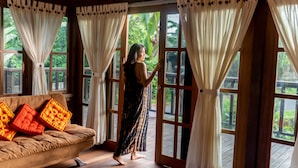 A room with a wooden floor, a brown sofa with orange cushions, and large windows with white curtains. - Villa Waturenggong Ubud (Ubud)