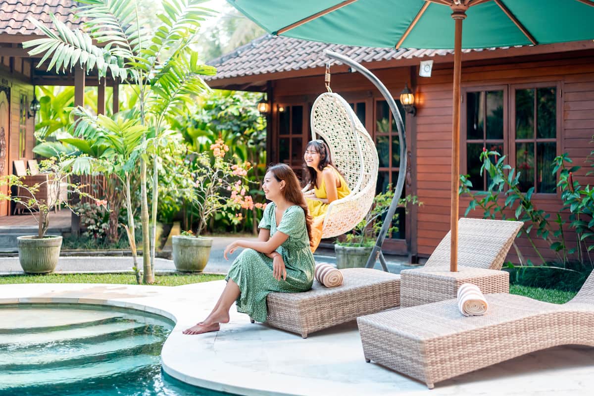 Dos sillones de mimbre junto a una piscina, con una casa de madera y abundante vegetación al fondo.