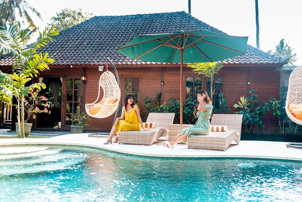 Un espace aménagé au bord de la piscine, avec des fauteuils de relaxation en osier, un fauteuil suspendu et un grand parasol.