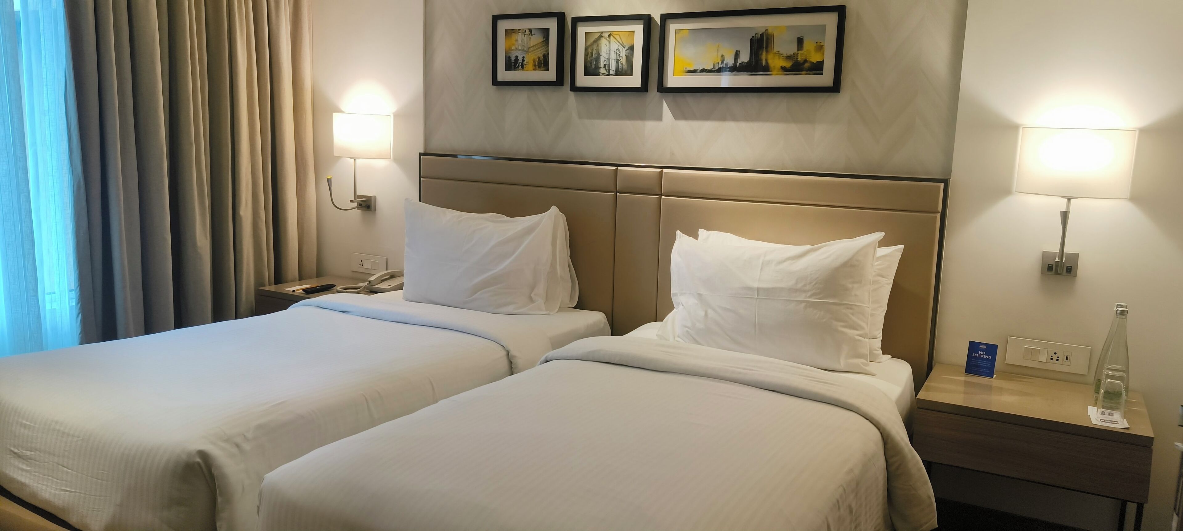 A hotel room with two beds, a nightstand with a lamp, a phone, and a small table with a bottle and a sign.