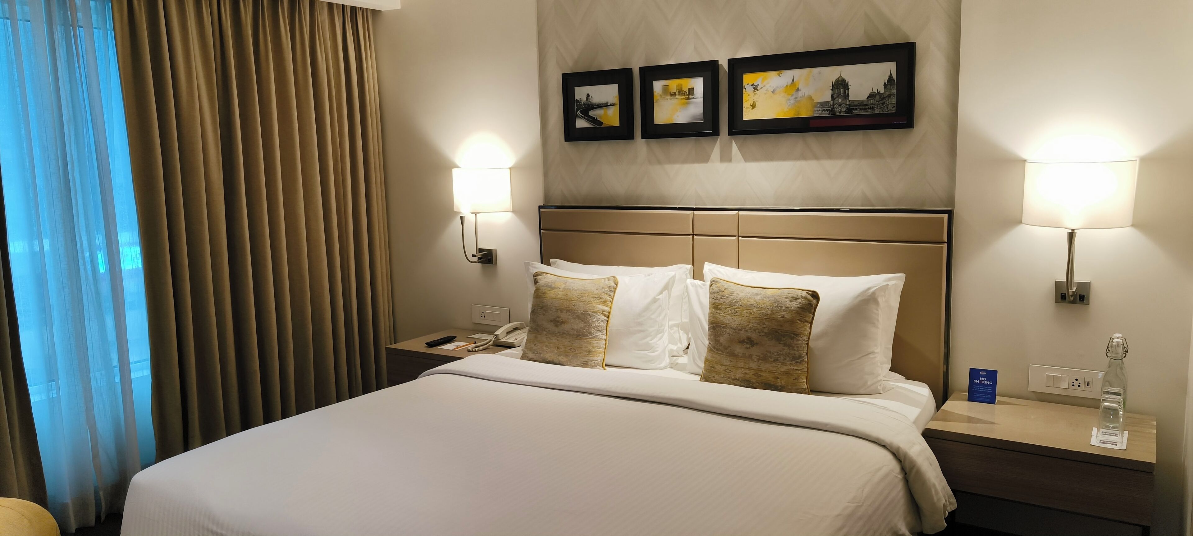 A hotel room with a large bed, two bedside lamps, a nightstand with a phone and a bottle, and three framed pictures on the wall.