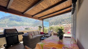 A covered outdoor patio with a wooden ceiling, a grill, wicker furniture, and a table with a colorful tablecloth.