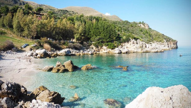 A coastal landscape with clear turquoise waters, rocky shorelines, and a hillside with dense greenery.