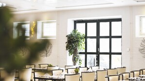 Un comedor moderno con mesas y sillas, ventanas amplias y una planta en maceta en el centro.