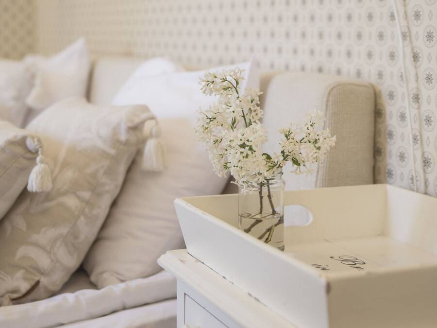 Una cama bien hecha con ropa blanca y almohadas, una mesita de noche con un florero y una lámpara en la pared.