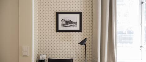 Une pièce avec un papier peint à motifs, un tableau encadré au mur, un bureau avec une lampe et une chaise.