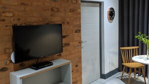 A modern living room with a brick accent wall, a flat-screen TV on a white shelf, a dining area with a table and chairs, and a clock on the wall. - Junction 502 Apartments (Pretoria)