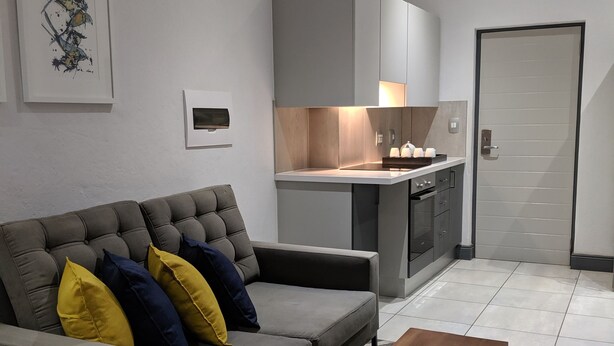 A modern living room with a grey sofa, a wooden coffee table, and a kitchen area with grey cabinets and a built-in oven. - Junction 502 Apartments (Pretoria)