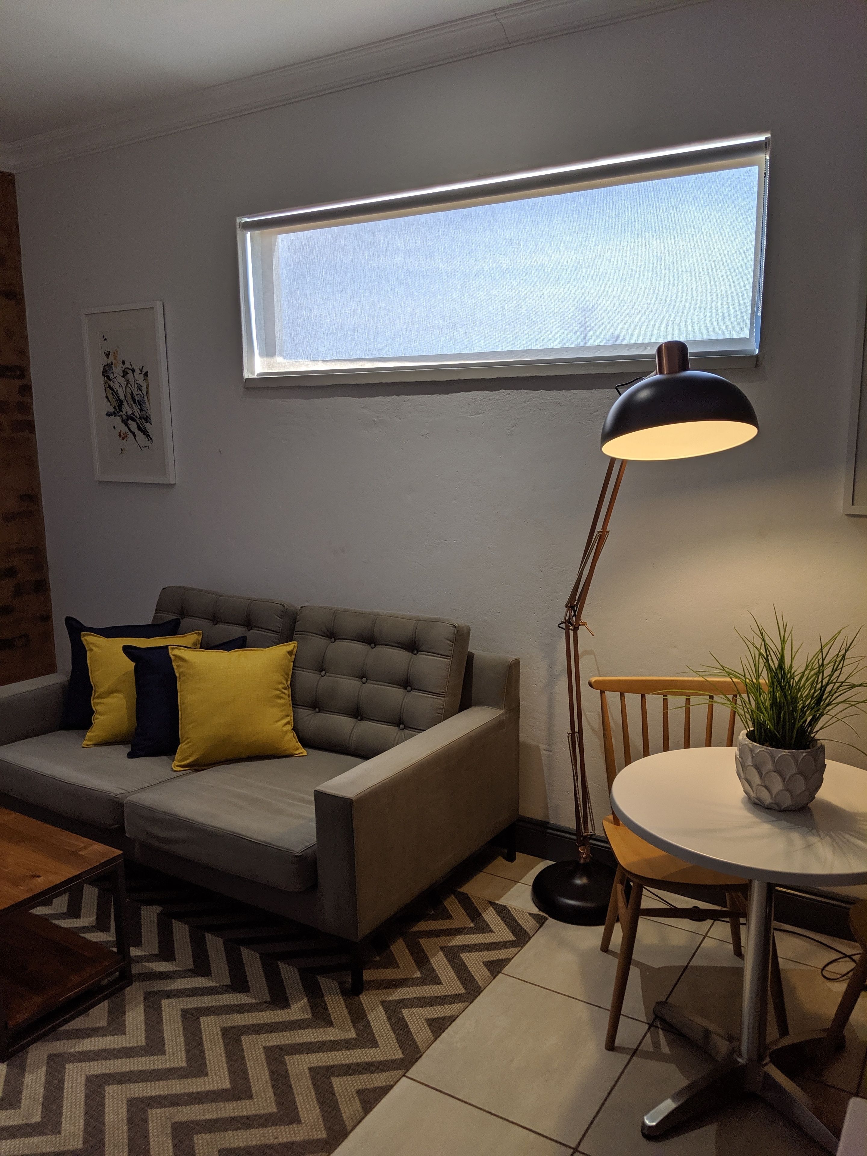A living room with a grey sofa, yellow cushions, a round table with a potted plant, a floor lamp, and a window with blinds.