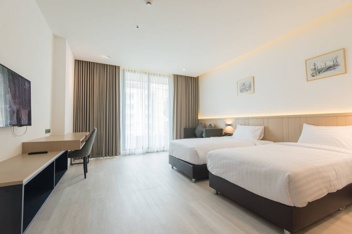 Habitación de hotel con escritorio, dos camas, televisor y un amplio ventanal con cortinas.
