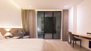 A modern hotel room with a large bed, a desk, and a chair. There is a sliding glass door leading to a balcony with a green wall. - Madison Bangkok (Bangkok)