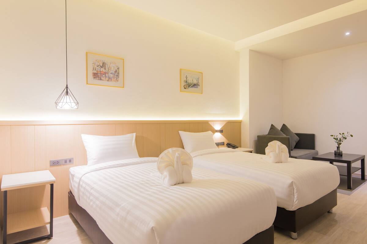 Habitación de hotel con dos camas, una mesita de noche, un escritorio, una silla, una lámpara y dos cuadros en la pared.