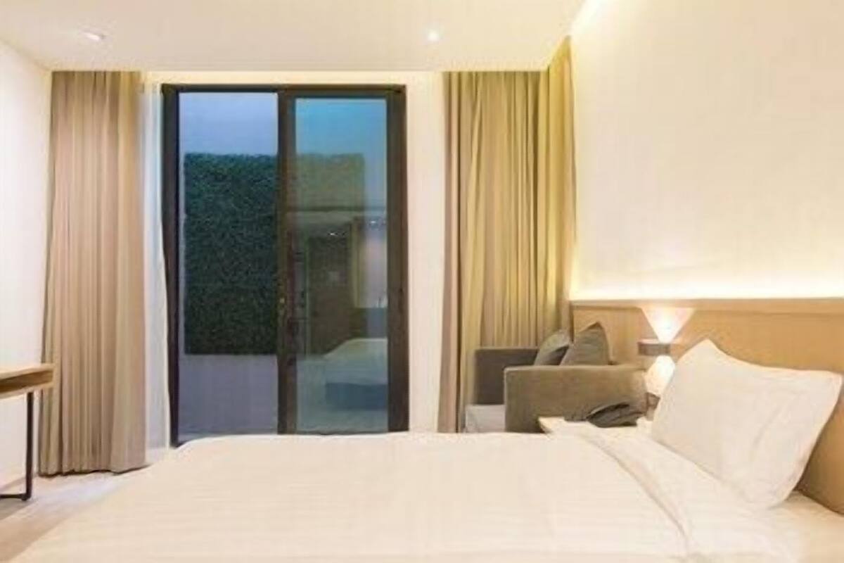 Habitación de hotel con cama, mesita de noche, lámpara, silla y puerta corrediza de vidrio que da a un balcón.