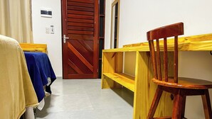 Habitación de hotel con puerta de madera, cama con cubierta color beige y azul, una silla de madera y un banco con cubierta amarilla.