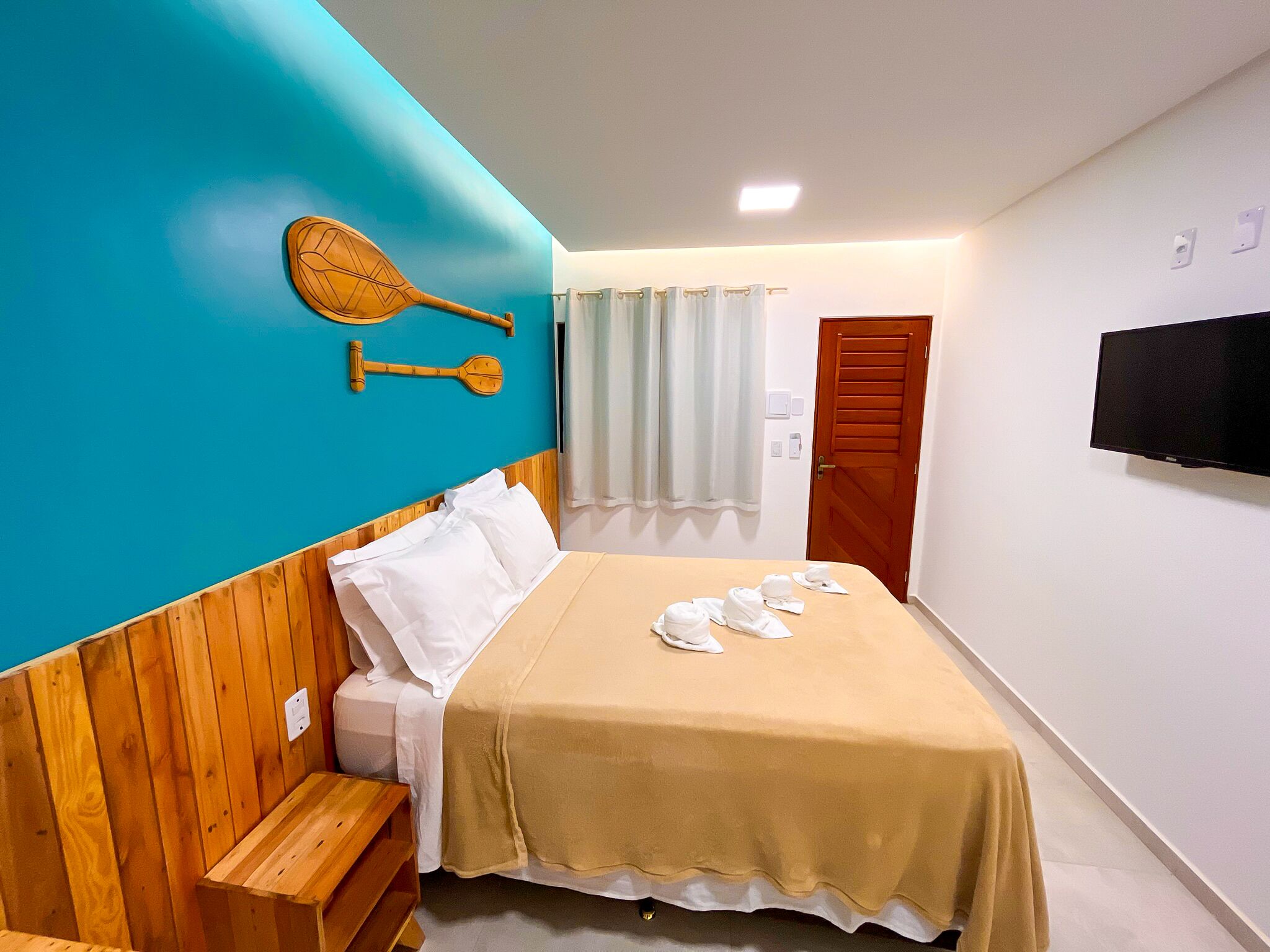 Habitación de hotel con cama, cabecero de madera, televisor montado en la pared y ventana con cortinas.