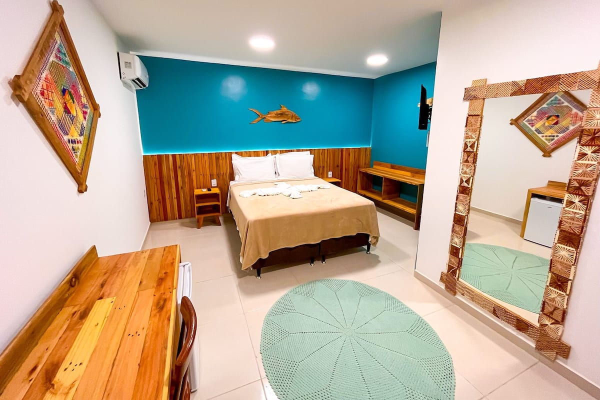 A compact hotel room with a bed, a small wooden table, a mirror, and a wall-mounted air conditioner.