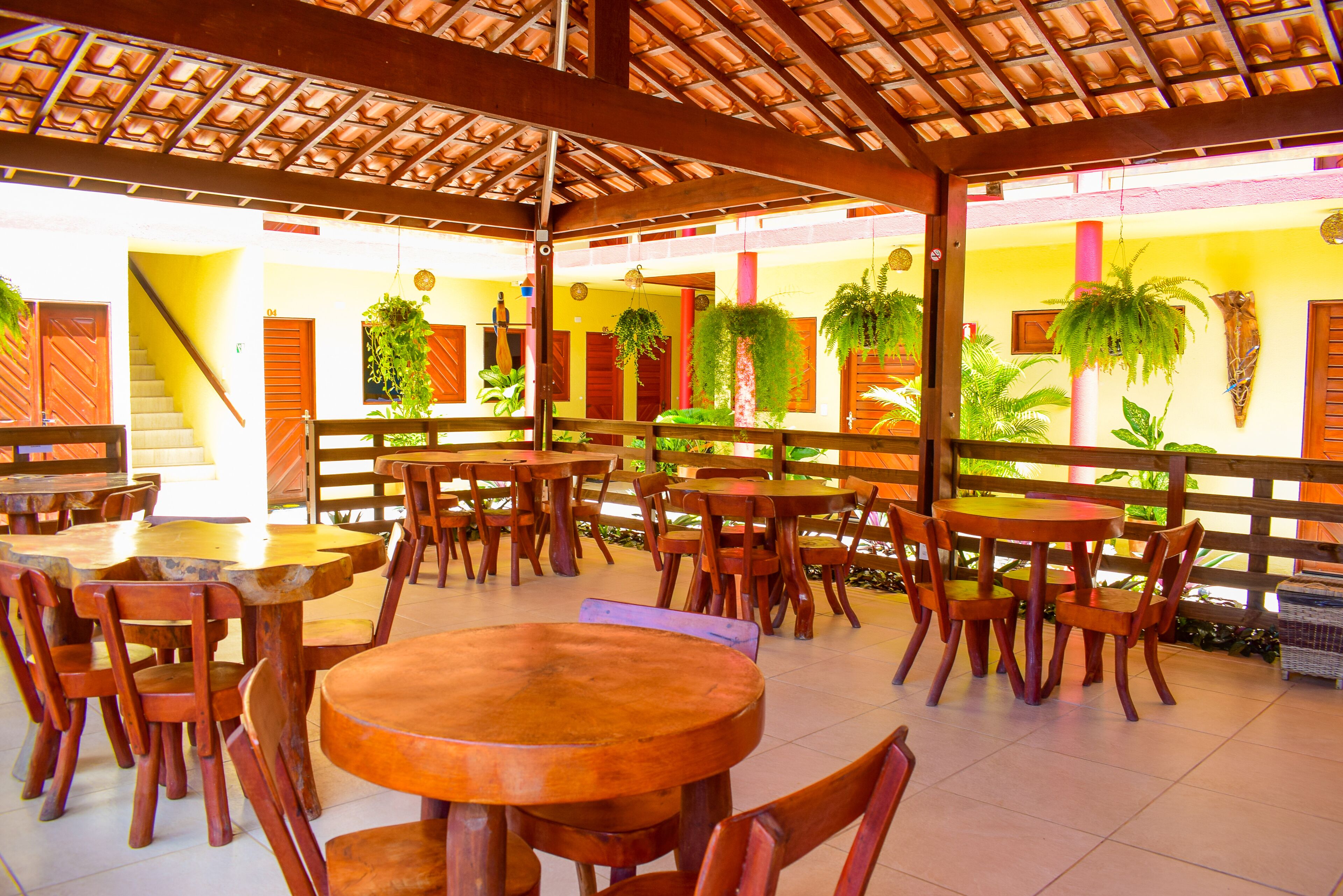 An outdoor cafe with wooden tables and chairs, surrounded by potted plants and a tiled floor.