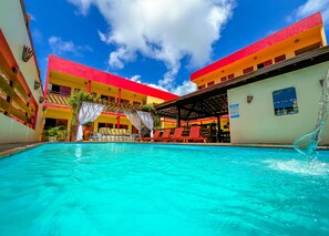 Una piscina con agua azul cristalina, rodeada de edificios con fachadas rojas y amarillas, y una zona de estar cubierta con sillones reclinables rojos.