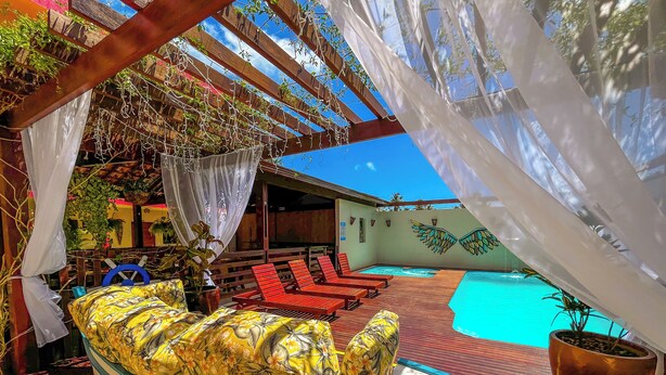Un área junto a la piscina con sillas reclinables rojas, una zona de estar techada con cojines florales amarillos, y un cielo azul despejado.