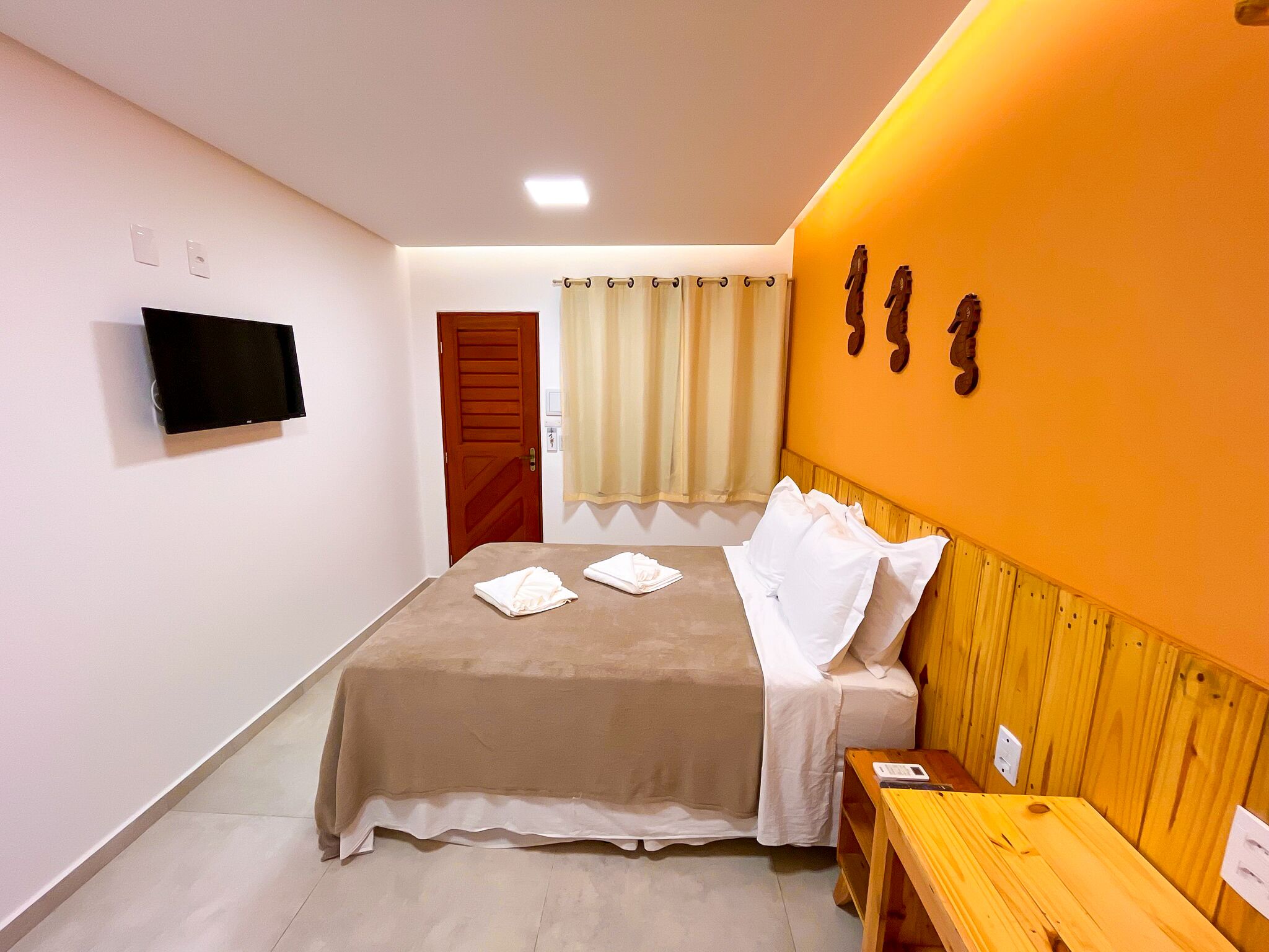 A hotel room with a bed, a wooden bench, a TV mounted on the wall, and a window with curtains.