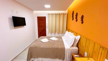A hotel room with a bed, a wooden bench, a TV mounted on the wall, and a window with curtains.